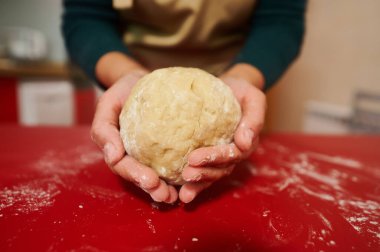 Taze hamuru tutan ve pişirmeye hazır ellerin yakın çekimi. Kırmızı yüzey ve un sıcak, kırsal bir his yaratır. Yemek ve yemek temaları için idealdir..