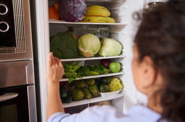 Bir kadın taze, renkli meyve ve sebzelerle dolu bir buzdolabını açıyor. Sahne sağlıklı beslenme ve canlı yaşam tarzı seçimlerini vurguluyor, çeşitli ürünlere odaklanarak..