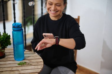 Gülümseyen bir kadın akıllı telefonunu tutarken akıllı saatini kontrol eder. Ahşap bir masada mavi bir su şişesinin yanında oturuyor ve rahat bir atmosfer yaratıyor. Teknoloji ve yaşam tarzı temaları için mükemmel.