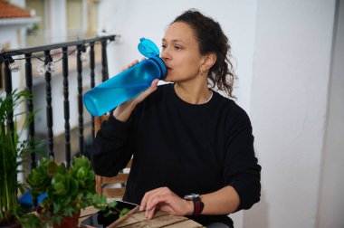 A woman sips water from a blue bottle on her balcony, surrounded by potted plants, capturing a serene moment of relaxation and hydration. clipart