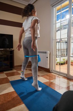 A young girl engages in a fitness routine indoors, using a blue yoga mat and resistance band. The sunlight streaming through the window creates a peaceful workout environment. clipart