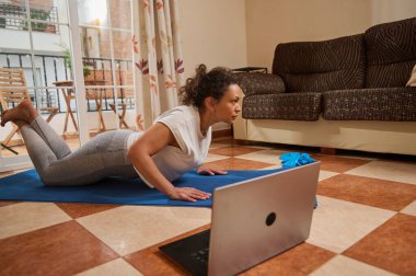 Bilgisayarındaki çalışmanın ardından, bir kadın evdeki minderde yoga pozu veriyor. Konforlu oturma odası, spor yapmak için huzurlu ve odaklanmış bir atmosferi ön plana çıkarıyor..