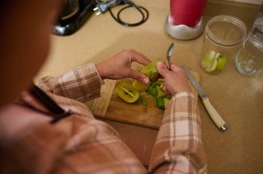 Tahta bir tahtada kivi soyup kesen, besleyici bir meyve püresi hazırlayan birinin yakın çekimi. Kurulum arka planda görünür bir blender ve diğer meyveleri içerir.