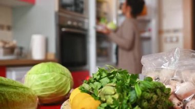 Ön planda, marul, limon, maydanoz ve Brüksel lahanası da dahil olmak üzere canlı bir taze sebze koleksiyonu bulunuyor. Arka planda bir kişi bu rahat dolaptan eşyalar alıyor.