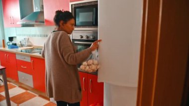 Bir kadın modern bir mutfakta, parlak kırmızı dolaplarıyla eşyaları düzenliyor. İşlevsel ve şık mutfak düzenini vurgulayarak mekanı düzenlemeye odaklanmış görünüyor..