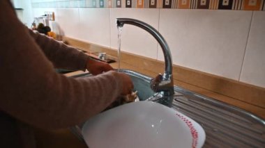 Mutfak lavabosunda akan suyun altında sebze yıkayan yakın plan bir el. Sahne, mutfak ortamının sıcaklığını ve umutsuzluğunu, temizliği ve sağlıklı bir yaşam tarzını yansıtıyor..