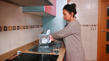 Bir kadın mutfak lavabosunda temiz bir sürahi dolduruyor. Modern mutfakta döşeme ve ahşap dolaplar var. Bu sahne günlük rutinleri ve ev verimliliğini vurguluyor..