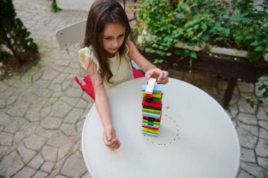 A young girl intently focuses on stacking a tower of colorful blocks at an outdoor table. The scene captures a moment of concentration and creativity, surrounded by greenery and open space. clipart