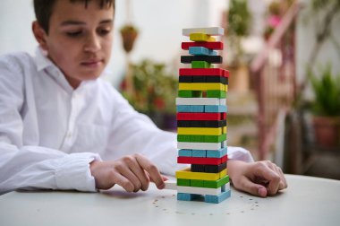 A boy in a white shirt concentrates on a colorful stacking game, balancing each piece delicately. The outdoor setting adds a natural backdrop, highlighting focus and precision in the playful activity. clipart