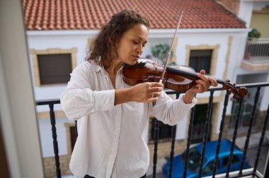 Odaklanmış bir kadın, kırmızı kiremitli çatıların ve konut binalarının büyüleyici zeminine karşı ustalıkla kemanını bir balkonda çalar. Huzurlu atmosfer onun müzik tutkusunu vurguluyor..