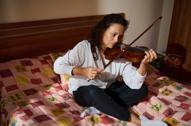 Gündelik bir ortamda, renkli bir yatağa oturmuş keman çalan yetişkin bir kadın. Doğal ışıklandırma odak noktasını ve odanın rahat atmosferini vurgular..