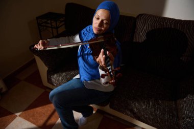 A woman wearing a blue hijab skillfully plays the violin while seated on a sofa, surrounded by the warm ambiance of her home. clipart