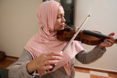 Pembe tesettürlü bir kadın evde ustalıkla keman çalıyor. Odaklanma ve zarafetini gösteriyor..