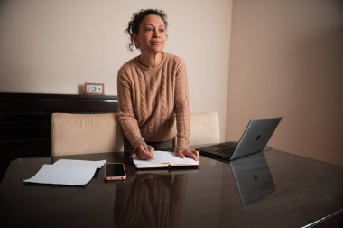Rahat bir kazak giyen bir kadın, evde çalışırken defterine yazıyor. Kurulumda bir dizüstü bilgisayar ve modern bir masadaki telefon da var..