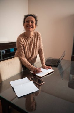 Neşeli bir kadın dizüstü bilgisayarı yakınlarda çalışırken not defterine not alır..