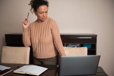 Sıcak süveter giymiş bir kadın, masasında düşünceli bir şekilde duruyor, evde çalışmak için dizüstü bilgisayar ve kalem kullanıyor..
