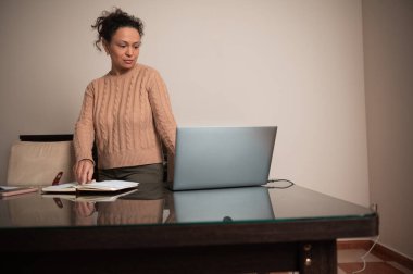 Bir kadın açık bir defterden bahsederken, bir dizüstü bilgisayar kullanarak masasında duruyor. Ayarlar ev ofisi ya da uzaktan çalışma ortamı olduğunu gösteriyor. Odaklanmış ve profesyonel görünüyor..