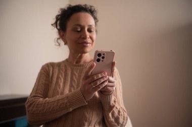 Rahat bir kazak giyen bir kadın evde akıllı telefonunu kullanmaktan hoşlanır. İçerde iyi aydınlatılmış bir ortamda cihazına odaklanmış ve rahatlamış görünüyor..