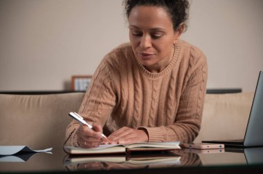 Bir kadın, dizüstü bilgisayarıyla evde uzaktan çalışarak bir deftere yazmaya odaklanır. Sıcak ortam onun konsantrasyonunu ve verimliliğini arttırıyor..