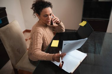 A woman is multitasking at her home office, using a phone and laptop, while reviewing documents. The setup reflects a modern work-from-home environment. clipart