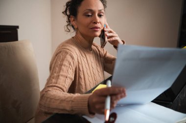 Ofisindeki bir telefon görüşmesi sırasında belgelere bakan odaklanmış bir kadın. Kurulum, modern iş yaşamında çoklu görevlerin vurgulandığı ev ortamının profesyonel bir çalışma olduğunu gösteriyor..