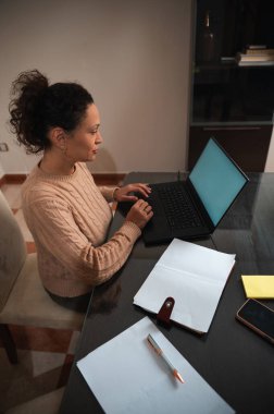 Odaklanmış bir kadın evdeki bilgisayarında çalışıyor, etrafı kağıt ve kalemle çevrili. Uzak çalışma, konsantrasyon ve üretkenlik kavramları için ideal.