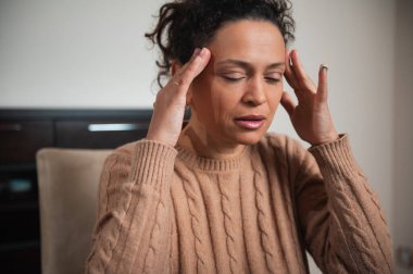 Kahverengi kazaklı bir kadın şakaklarını tutar, evde stres ya da rahatsızlık yaratır..