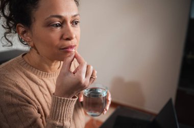 Düşünceli bir kadın evinde dizüstü bilgisayarının yanında otururken suyla birlikte bir hap alıyor. Ortam, ev çalışma ortamı içinde günlük sağlık ve sağlık rutinlerini ortaya koyuyor.