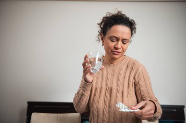 Rahat bir kazak giyen bir kadın, elinde bir bardak su tutarken hapları inceler, sağlık ve sağlık temalarını iletir.