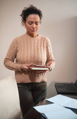 Bir kadın ev ofisinde elinde bir kitapla dizüstü bilgisayarın yanında duruyor. Ortam rahat ve sıcak, rahat bir ortamda odaklanmayı ve üretkenliği öneriyor..