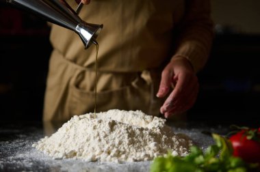 Close-up of a chef in an apron pouring oil onto a mound of flour, beginning the process of making fresh dough. Fresh ingredients are arranged in the foreground, indicating a vibrant and natural culinary experience. clipart