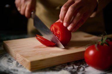 Bir insan taze yemek hazırlığını vurgulamak için tahtada sulu kırmızı bir domates dilimliyor. Görüntü, mutfak ortamında yapılan ev yapımı mutfak tatbikatlarının özünü yansıtıyor..
