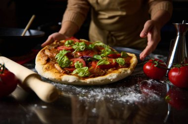 Bir şef taze domates ve fesleğenli ev yapımı pizza hazırlar ve geleneksel İtalyan mutfağının özünü yakalar. Konforlu mutfak dekorasyonu otantik yemek pişirme deneyimini geliştirir..