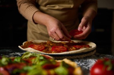 Pizzanın üzerine domates dilimleri yerleştiren, taze malzemeler sergileyen ve ev yapımı yemek pişirme sanatını sergileyen ellerin yakın çekimi..