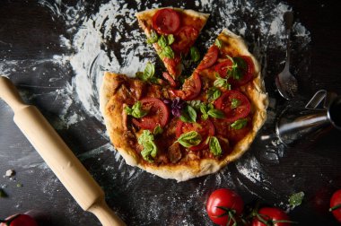 Taze domatesli ve fesleğenli lezzetli bir ev yapımı pizza, köy masasında. Un ve merdane otantik bir dokunuş ekler, İtalyan mutfağının özünü yakalar..