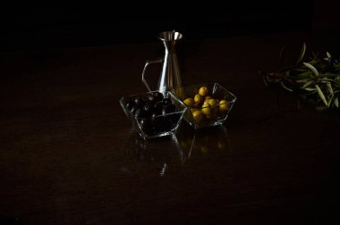 Koyu ahşap bir yüzey üzerinde metal yağ sürahisi bulunan cam kaselerde siyah ve yeşil zeytinlerin yer aldığı sofistike bir düzenleme..