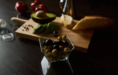 A selection of fresh olives and avocado slices on a cutting board with bread and tomatoes. Prepared for a healthy snack or appetizer on a dark, moody kitchen surface. clipart