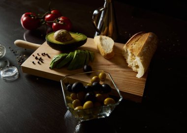 A rustic kitchen setup with sliced avocado, tomatoes, olives, and bread on a wooden board. Ideal for cooking, food styling, and healthy eating concepts in a Mediterranean atmosphere. clipart