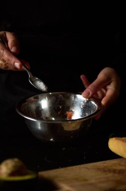 Metal bir kaseye tuz eklemek için yakın plan çekimde, bastırılmış ışıkta aşçılık hazırlığını vurguluyor. Görüntü, yakınlarda görünen malzemelerle yemek pişirmede odaklanma ve hassasiyeti vurgular.