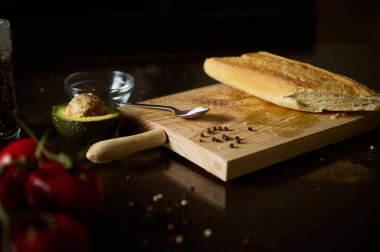 Koyu ahşap bir masa üzerinde taze ekmek, avokado ve domateslerin yer aldığı kırsal bir düzenleme. Organik gıda, aşçılık ya da kırsal mutfak temaları için ideal..