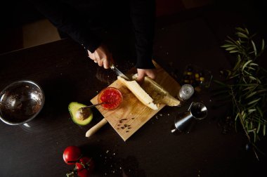 Bir insan, avokado, domates ve malzemelerle çevrili ahşap bir tahtanın üzerinde taze ekmek dilimleyip sade ve rahat bir mutfak atmosferi yaratır..