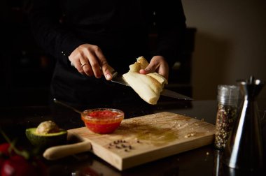 A person slices fresh bread on a wooden cutting board in a warm, inviting kitchen. Ingredients like avocado, spices, and sauce highlight a culinary atmosphere focused on simplicity and comfort. clipart