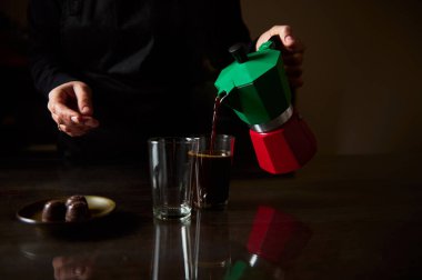 A person pours fresh coffee from a colorful moka pot into a glass, accompanied by chocolates on a table. clipart