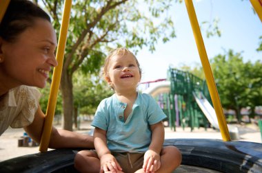 Neşeli bir çocuk, neşeli bir oyun parkında gülümseyen bir bakıcı ile oyun zamanı geçirir, güneşin tadını çıkarır..