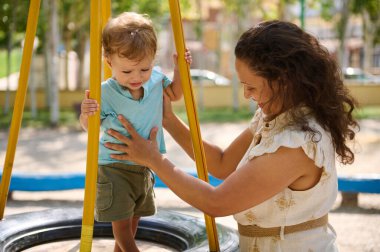 Şefkatli bir anne çocuğunun güneşli bir parkta salıncakta sallanmasına yardım eder..