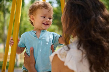 Sevimli çocuk neşeli bir şekilde sırıtıyor. Hem de canlı bir çocuk parkında annesi tarafından tutulurken. Sahne, canlı ve güneşli bir günle çevrili, sıcaklıkla ve sevgiyle dolu neşeli bir anı yakalar..