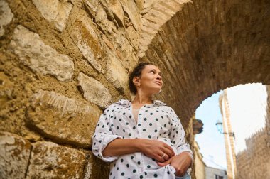 Puantiyeli bir kadın kemerin altındaki taş bir duvara dayanıyor ve tarihi bir yerde açık havada keşif yapıyor..