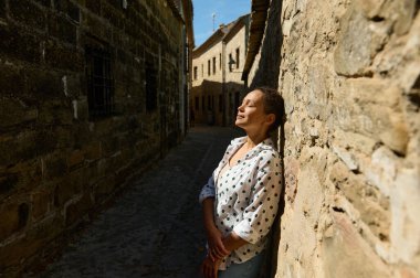 A woman leans against a stone wall, eyes closed, enjoying the warmth of the sun in a quaint, cobblestone alleyway. clipart