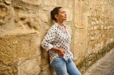 Bir kadın kırsal taştan bir duvara yaslanıyor, gözleri kapalı, güneşin tadını çıkarıyor. Sıradan bir polka bluz ve kot pantolon giyiyor, doğal bir ortamda rahatlık ve sükunet simgeliyor..