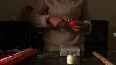 A person carefully wrapping a present on a table with red paper, a candle, and string while preparing for a festive occasion clipart